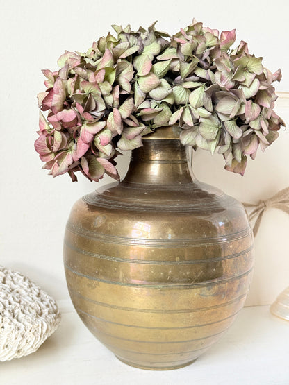 A vintage-style metallic gold vase with horizontal ridges, filled with dried pink and green hydrangea flowers, set against a neutral off-white background.