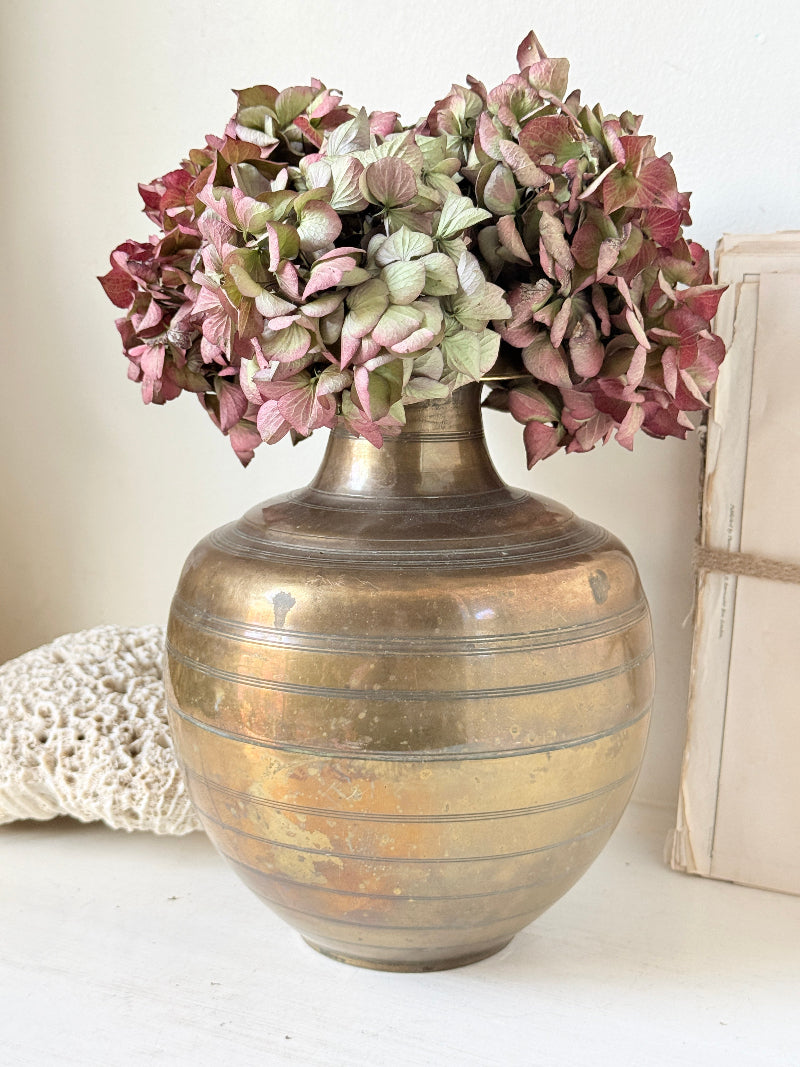 A vintage-style metallic gold vase with horizontal ridges, filled with dried pink and green hydrangea flowers, styled on a white surface next to a white crochet blanket and a beige box.