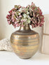 A vintage-style metallic gold vase with horizontal ridges, filled with dried pink and green hydrangea flowers, styled on a white surface next to a white crochet blanket and a beige box.