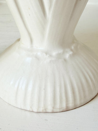 Close-up of a white ceramic vase base with a fluted, ribbed design and a narrow neck, set against a light off-white background.