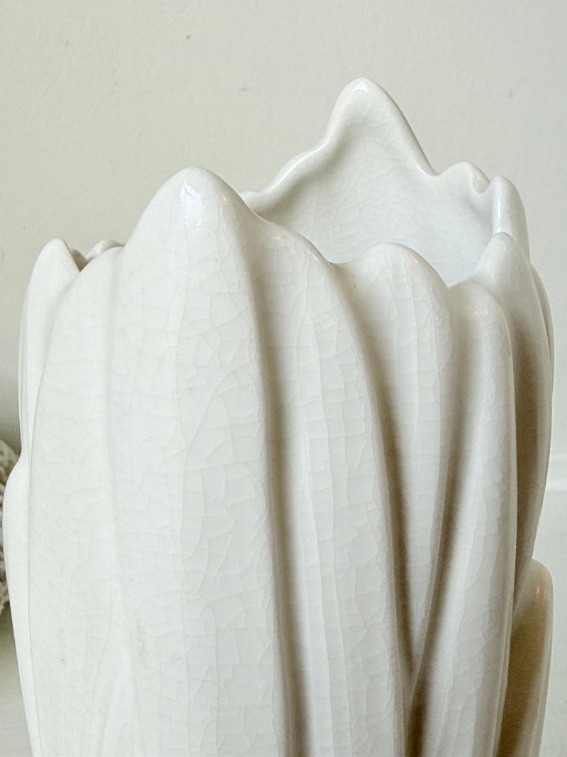 A close-up of a white ceramic vase with a textured, crackled glaze and a wavy, scalloped rim, set against a neutral beige background.