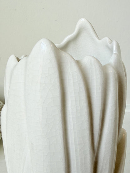 A close-up of a white ceramic vase with a textured, crackled glaze and a wavy, scalloped rim, set against a neutral beige background.