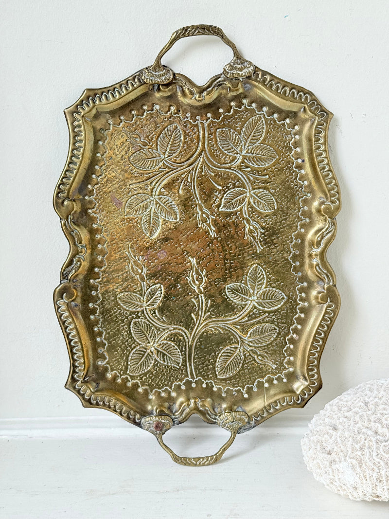 Vintage brass tray with embossed floral and leaf design, scalloped edge, and twin handles, photographed on a white surface