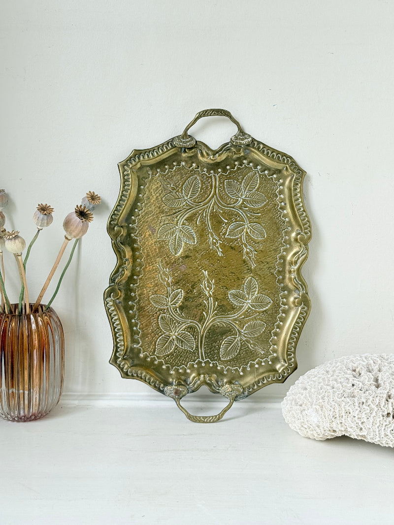 Vintage brass tray with embossed floral and leaf design, scalloped edge, and twin handles, photographed on a white surface
